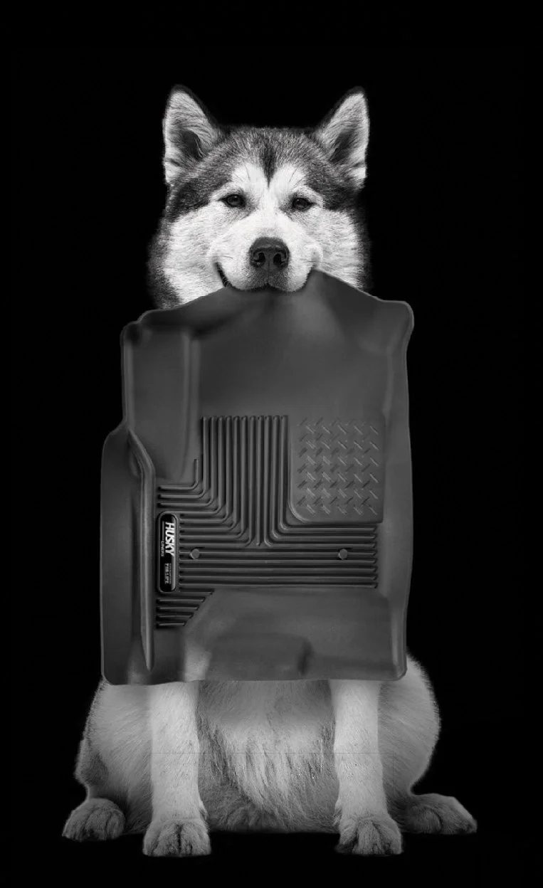 Husky Liners ad: a husky dog holds a black Husky floor liner in its mouth.